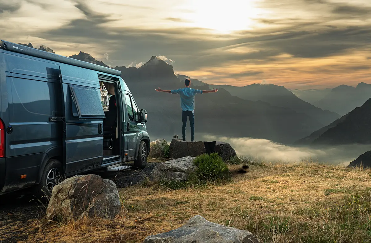 Ein Wohnmobilfahrer stehr neben seinem Wohnmobil mit ausgebreiteten Armen und schaut in die Berge